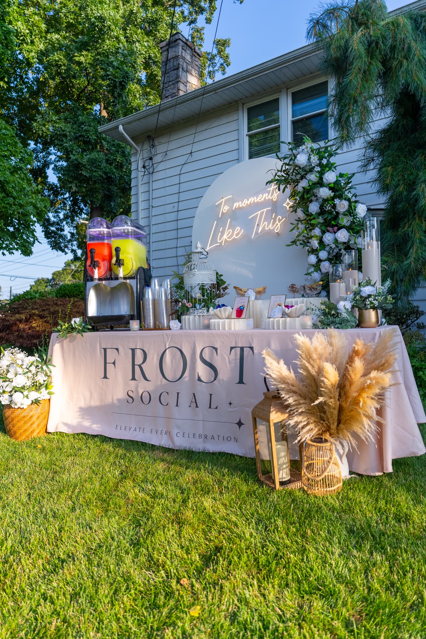 Golden-hour outdoor buildout with 'To moments Like This' neon arch, floral column, and pampas grass