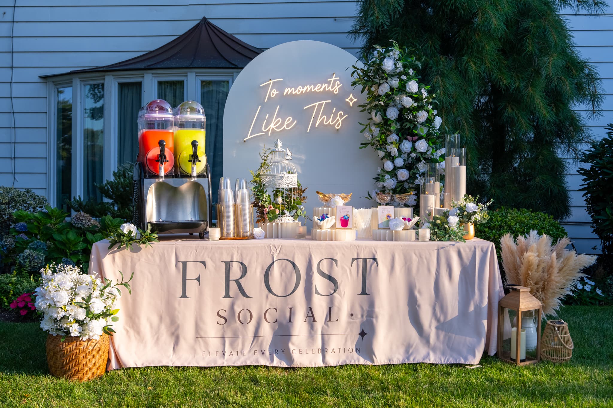 Evening buildout with lit neon arch, branded FROST SOCIAL linen, twin-barrel machine, and floral column