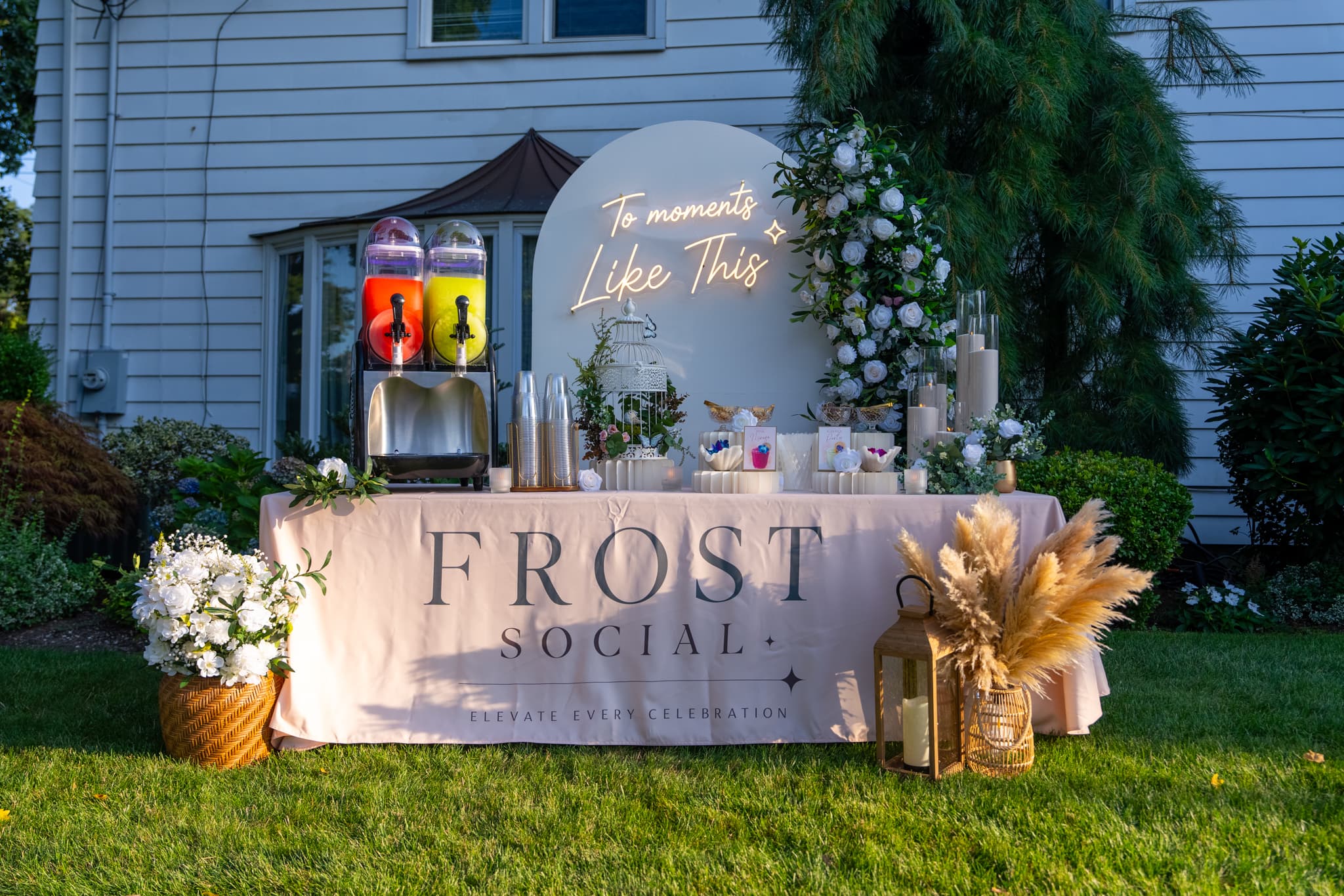Frost Social frozen mocktail bar buildout at golden hour: twin-barrel machine, floral arch with 'To moments Like This' neon, branded linen tablecloth